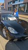 A woman in sunglasses, wearing a blue blazer and white shirt, stands beside a black sports car with a white sign in the window that reads “Hampton Grand Marshall.”
